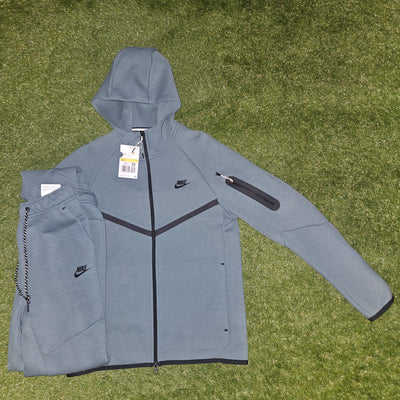A gray Nike hooded zip-up jacket laid flat on green grass, with matching gray joggers folded beside it. The jacket has black trim, a black Nike swoosh, and a black zip pocket on the upper sleeve.