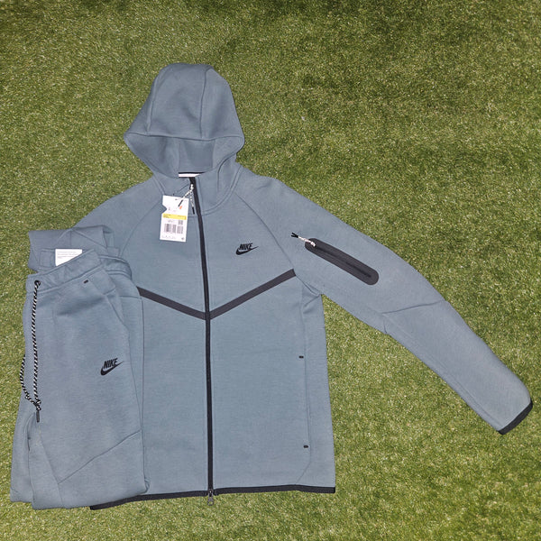A gray Nike hooded zip-up jacket laid flat on green grass, with matching gray joggers folded beside it. The jacket has black trim, a black Nike swoosh, and a black zip pocket on the upper sleeve.