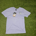 A light lavender short-sleeve t-shirt by The North Face, laid flat on green grass. The shirt features a small gray logo patch on the left chest and has its original hangtag and paper tag attached.
