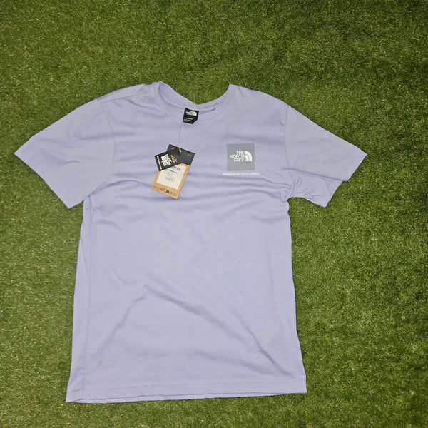A light lavender short-sleeve t-shirt by The North Face, laid flat on green grass. The shirt features a small gray logo patch on the left chest and has its original hangtag and paper tag attached.