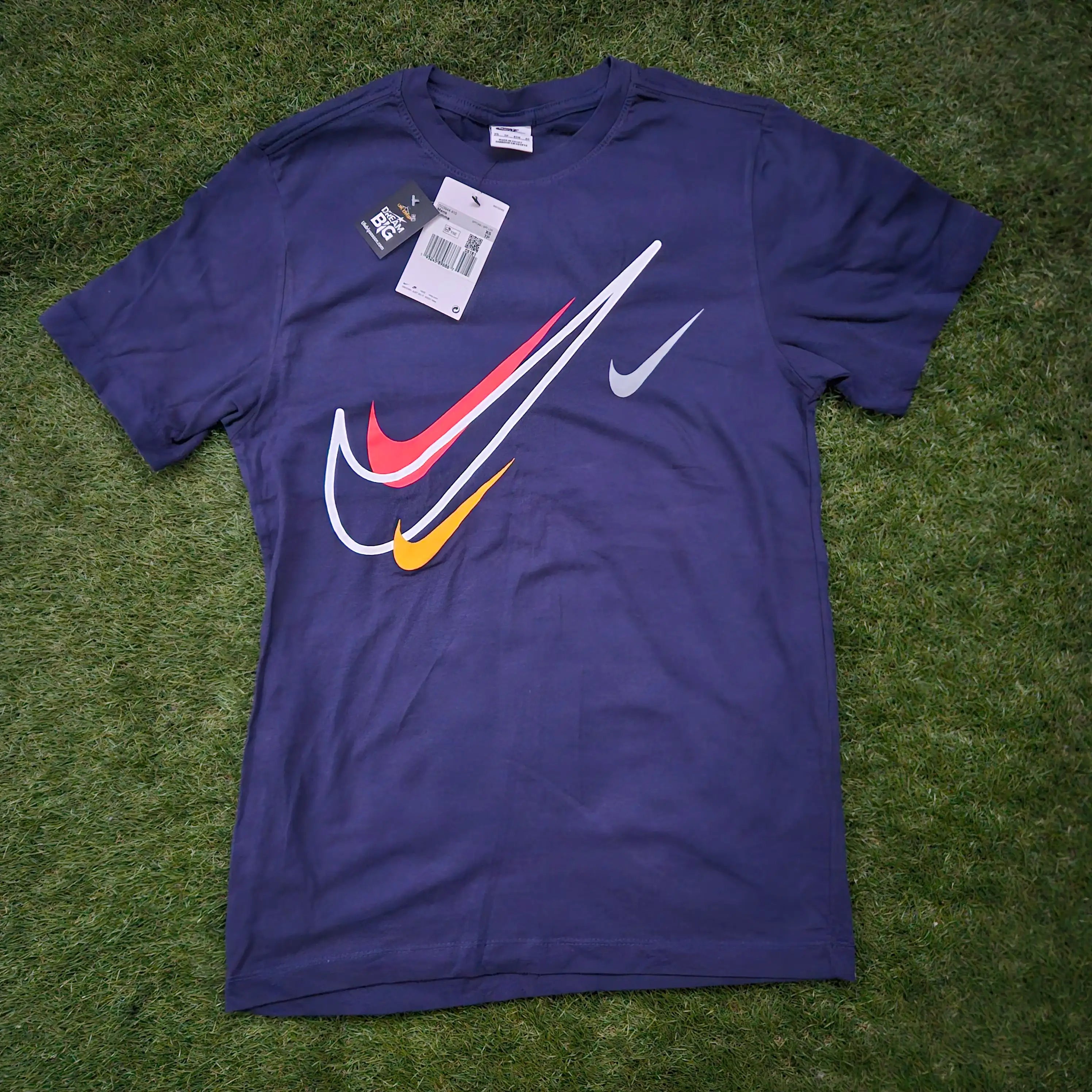 A Nike short-sleeve t-shirt in dark purple, laid flat on green grass. The shirt features a large graphic chest print with a white swoosh, a red swoosh, and a yellow swoosh layered over a white diagonal swoosh, plus a smaller white swoosh on the opposite chest.
