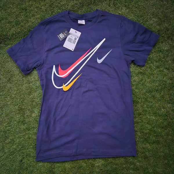 A Nike short-sleeve t-shirt in dark purple, laid flat on green grass. The shirt features a large graphic chest print with a white swoosh, a red swoosh, and a yellow swoosh layered over a white diagonal swoosh, plus a smaller white swoosh on the opposite chest.